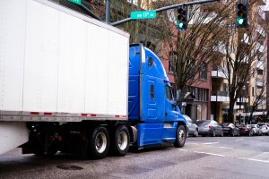 Big rig semi truck in blue with trailer moving by urban city street with modern building and cars