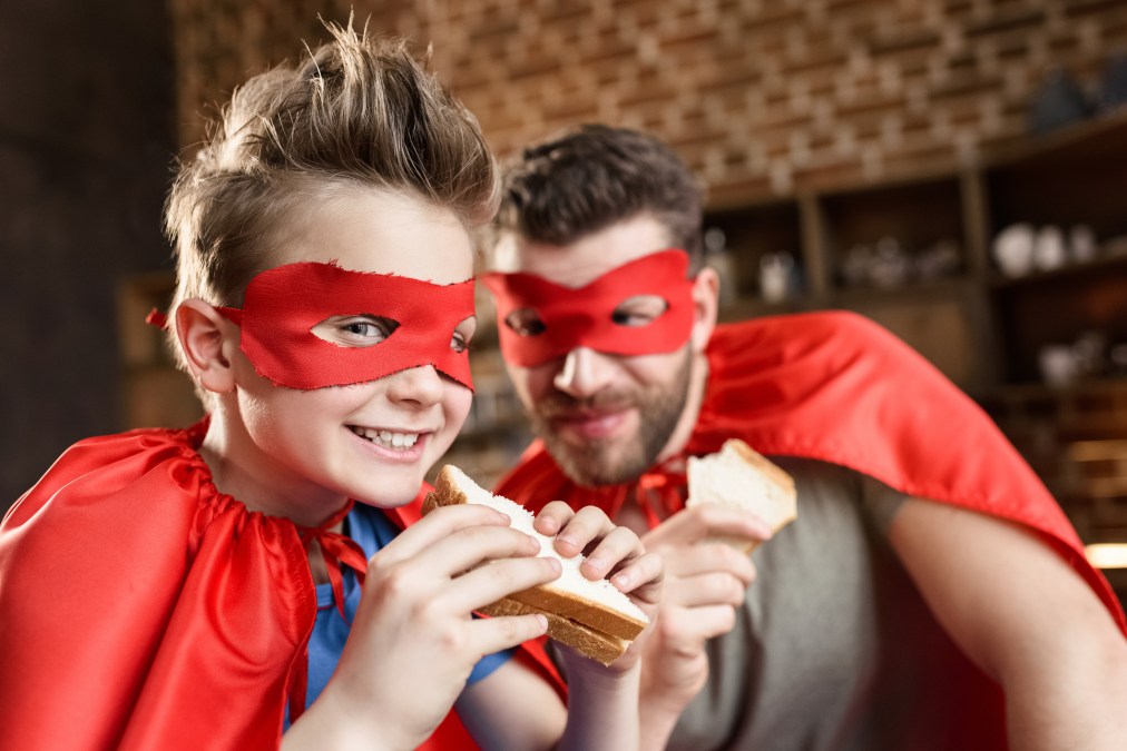 father and son in red superhero costumes eating sandwiches at home