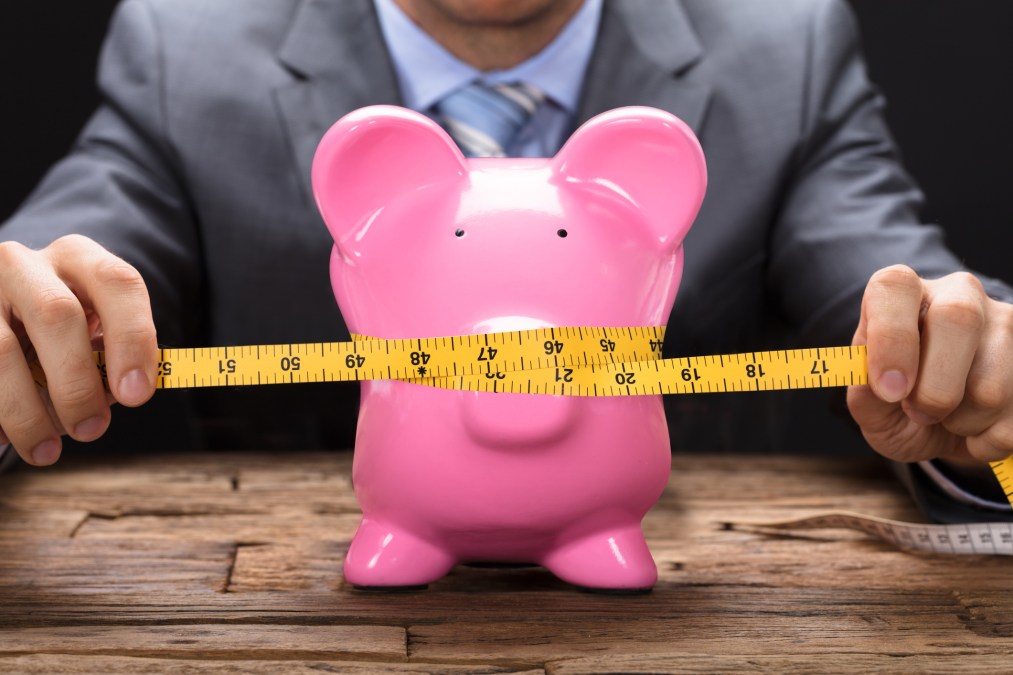 Businessman Squeezing Piggybank With Tape Measure On Table