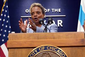 Chicago Mayor Lori Lightfoot photographed speaking at a wooden podium with a city seal in front of a sign saying "office of the mayor" and an American flag.