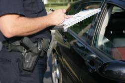 A photograph of a policeman writing on a clipboard while talking to someone seated in their car.