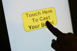 The silhouette of a finger is seen touching a button on a screen that says "touch here to cast your ballot"