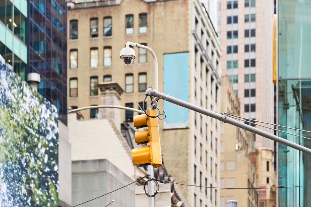 Security camera and traffic light are mounted on a pole, keeping watch over a bustling city street in New York City.