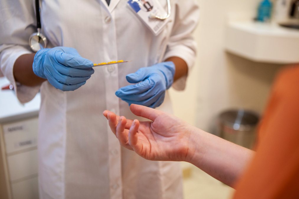 A person wearing orange holds out their hand to receive medication from a person wearing a white coat and blue nitrile gloves with a stethoscope around their neck.